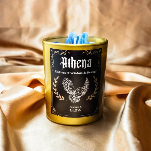 Złota świeca sojowa Athena marki Gloomy Glow z etykietą przedstawiającą sowę, ustawiona na eleganckim złotym materiale.