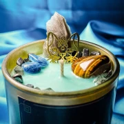 Detale świecy Bastet: turkusowy wosk sojowy, surowy kwarc dymny, tygrysie oko oraz złoty amulet w kształcie głowy kota.