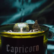 Zbliżenie na etykietę świecy Capricorn The Grounded ze złotym amuletem w kształcie góry i naturalnymi minerałami na powierzchni..webp