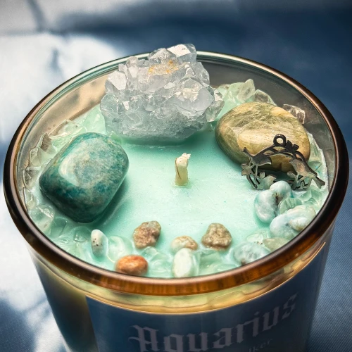 Widok z góry na jasnoniebieski wosk świecy zodiakalnej Aquarius od gloomandglow. Ezoteryczne świece sojowe ozdobione dużą szczotką kryształu, gładkim zielonym minerałem oraz metalową zawieszką.