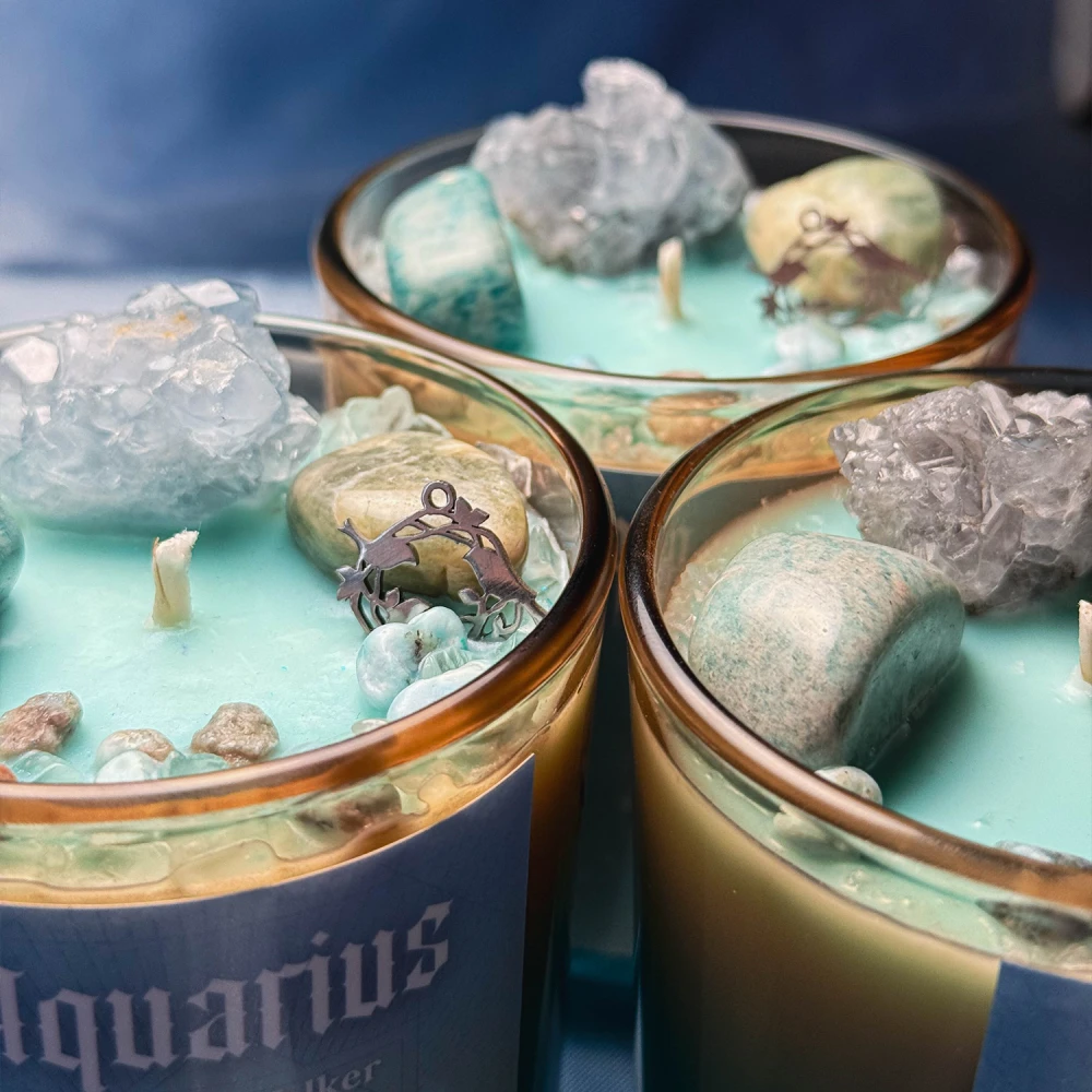 Zbliżenie na detale trzech ezoterycznych świec sojowych z serii zodiakalnej Aquarius od gloomandglow. Estetyczna kompozycja przedstawiająca różnorodne naturalne kryształy, drobne kamyczki i magiczne dekoracje.