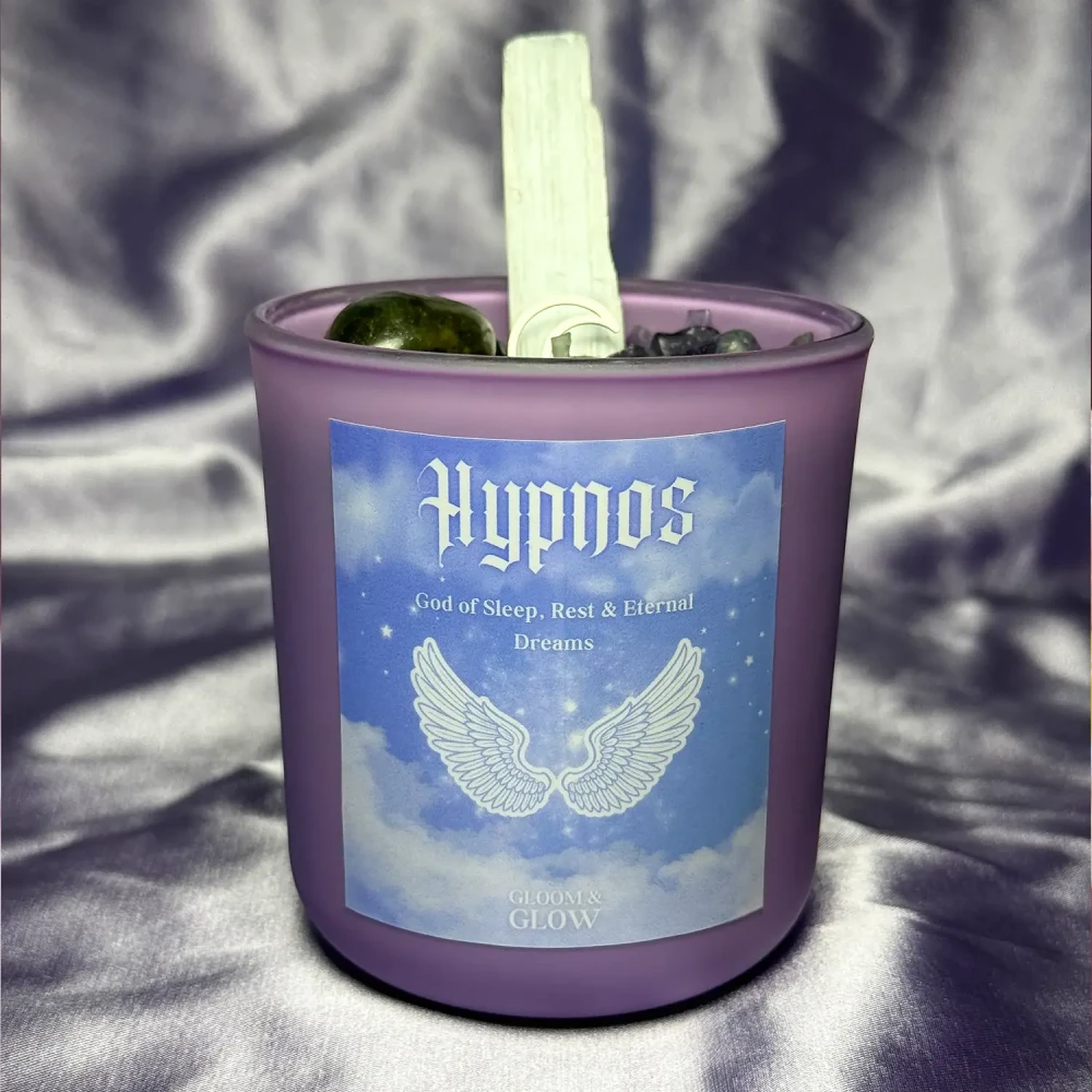 Fioletowa ezoteryczna świeca sojowa Hypnos marki Gloomandglow z etykietą ze skrzydłami i kryształami na satynowym tle.