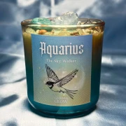 Turkusowa świeca w szkle z serii Aquarius (Wodnik) od gloomandglow. Te ezoteryczne świece sojowe mają jasną etykietę z grafiką lecącego ptaka i są zaprezentowane na błękitnym, satynowym materiale.