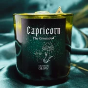 Ciemnozielona świeca sojowa dla znaku zodiaku Koziorożec marki Gloomy Glow, ustawiona na tle błyszczącej, szmaragdowej satyny..webp