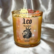 Bursztynowa świeca w szkle z serii zodiakalnej Leo (Lew) od gloomandglow. Te ezoteryczne świece sojowe mają ciepłą, pomarańczowo-złotą etykietę z lwem w koronie i stoją na jasnym, satynowym tle.