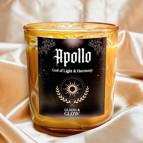 Świeca sojowa Gloomy Glow – Apollo God of Light w bursztynowym słoiku z czarną etykietą, otoczona elegancką, złotą tkaniną.